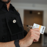 A person in a black uniform showcases an ID card connected to the Custom Compact Key-Bak Retractable Carabiner ID Badge Reel with Key Ring and Your Logo, enhancing durability.