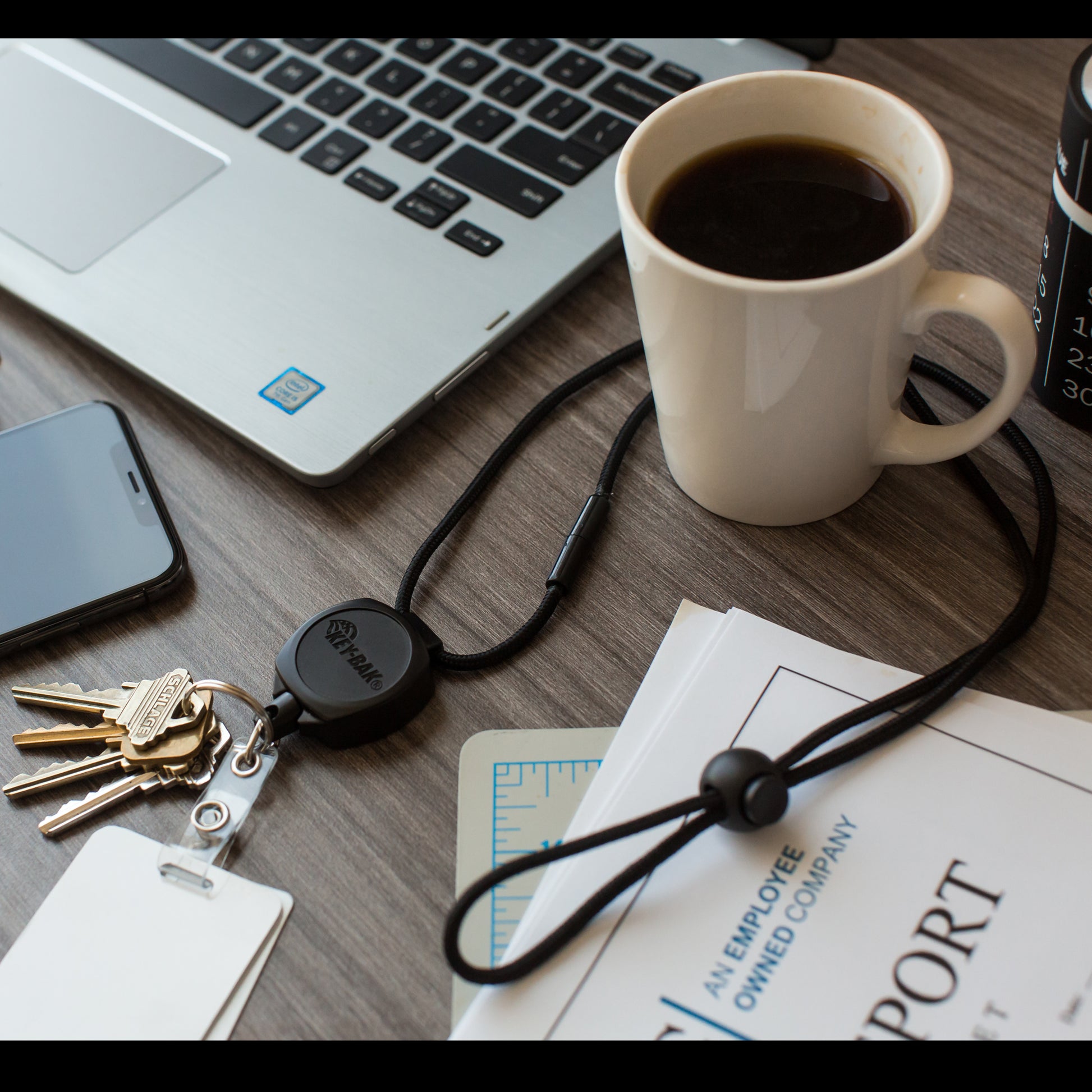 A desk with a laptop, smartphone, coffee cup, keys, and documents, including one labeled "AN EMPLOYEE OWNED COMPANY." A Key-Bak Heavy Duty Paracord Lanyard With Sidekick Retractable ID Badge & Key Reel (0KB1-0A44) is also present.