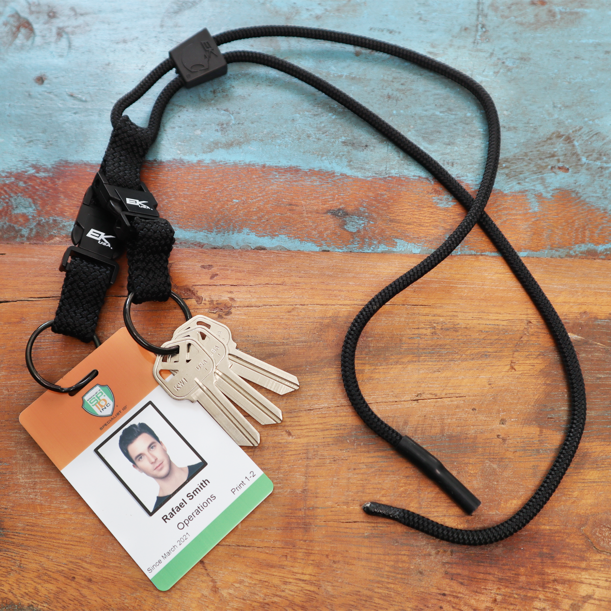 An EK Black Lanyard With Two Detachable Key Rings (10046) by EK USA, adorned with keys and a badge labeled "Rafael Smith, Operations, Plant 12," lies on a wooden surface.