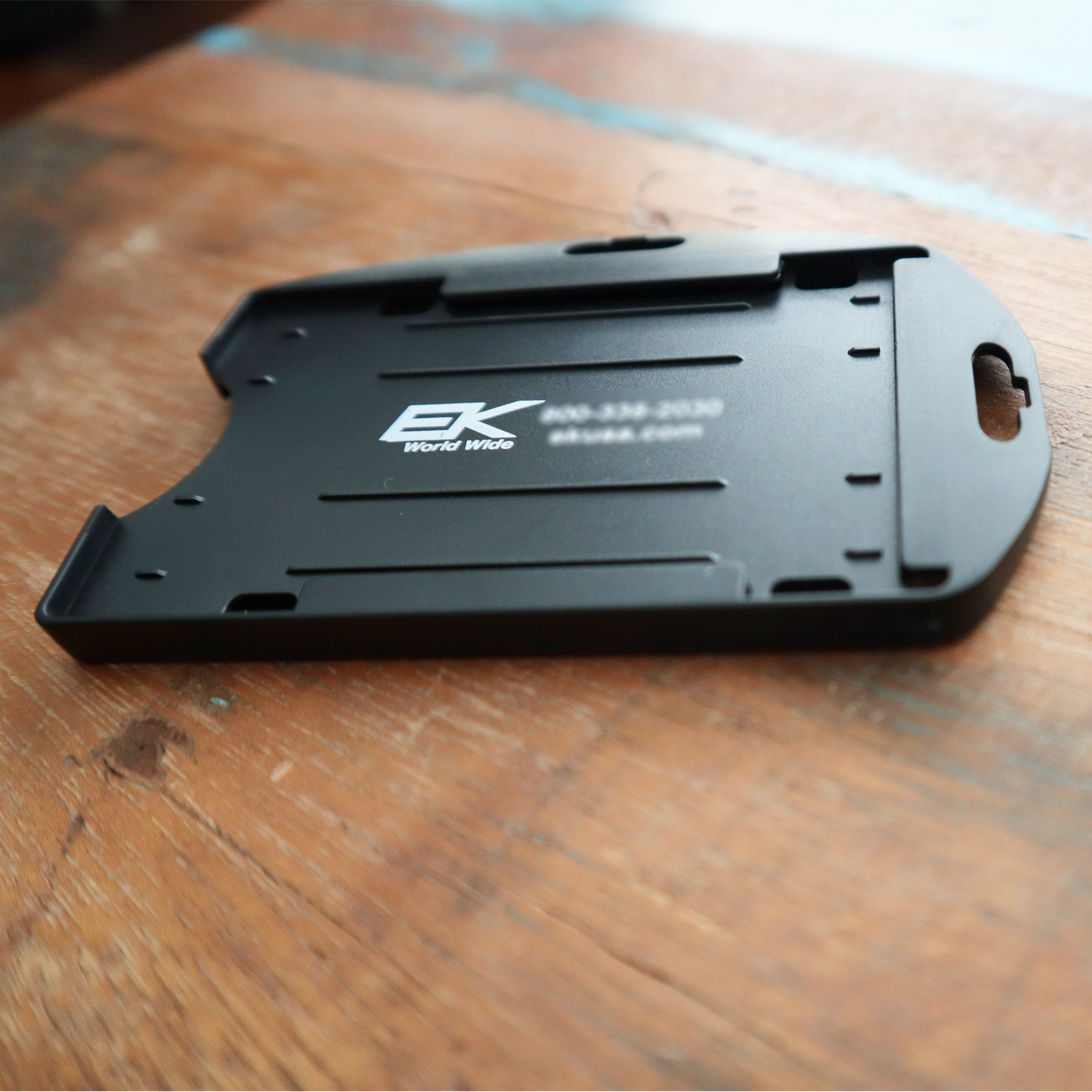 The Black EK Dual Sided Smart Card Holder with Slot Holes (10574) by EK USA features dual-sided black plastic construction with the company logo and contact information, elegantly displayed on a wooden surface to highlight its versatility as a smart card holder.