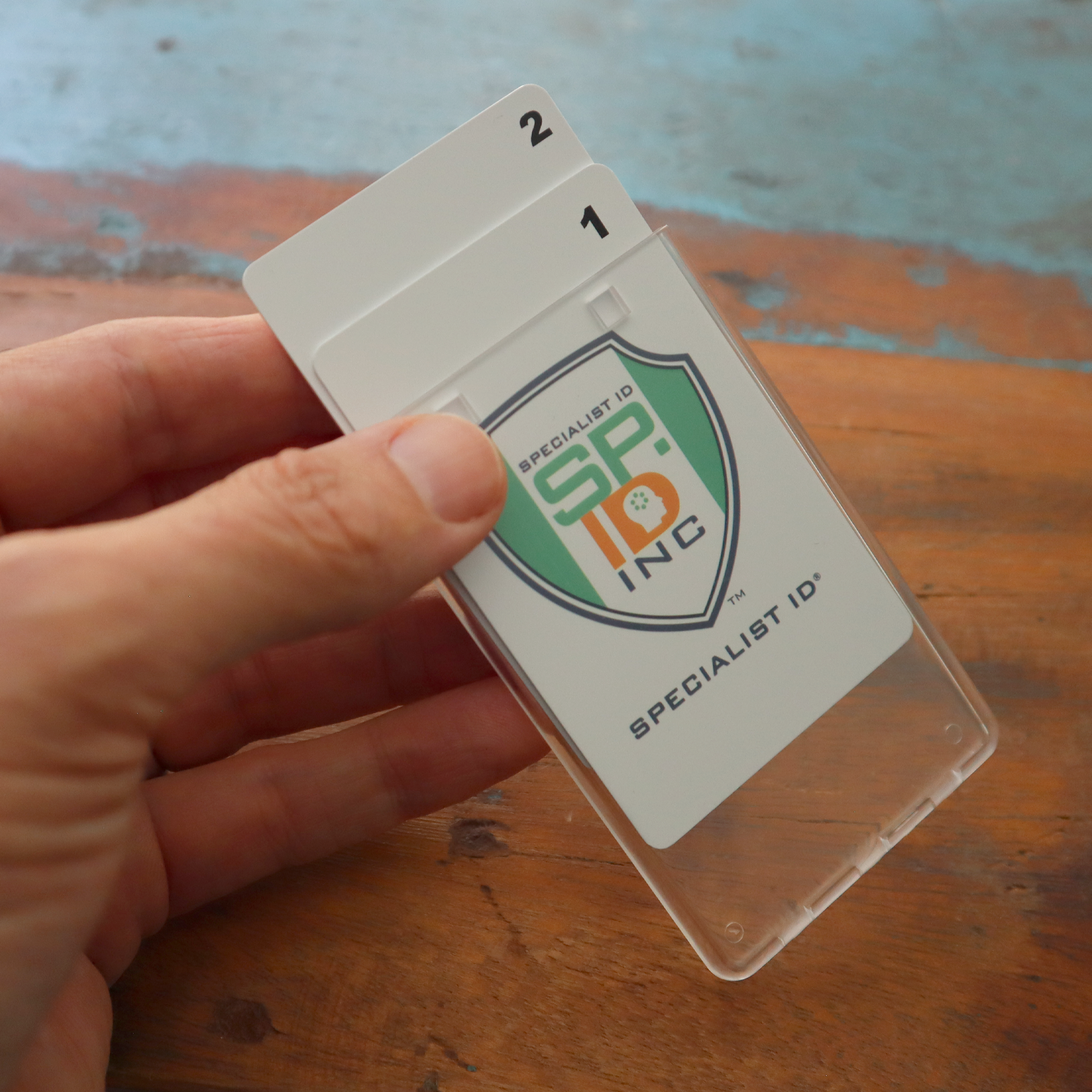 A hand holds a Clear Vertical Permanent Locking Plastic Card Holder (P/N 1840-6045) containing two cards labeled "1" and "2," featuring a logo reading "SP INC Specialist ID" on a wooden surface.