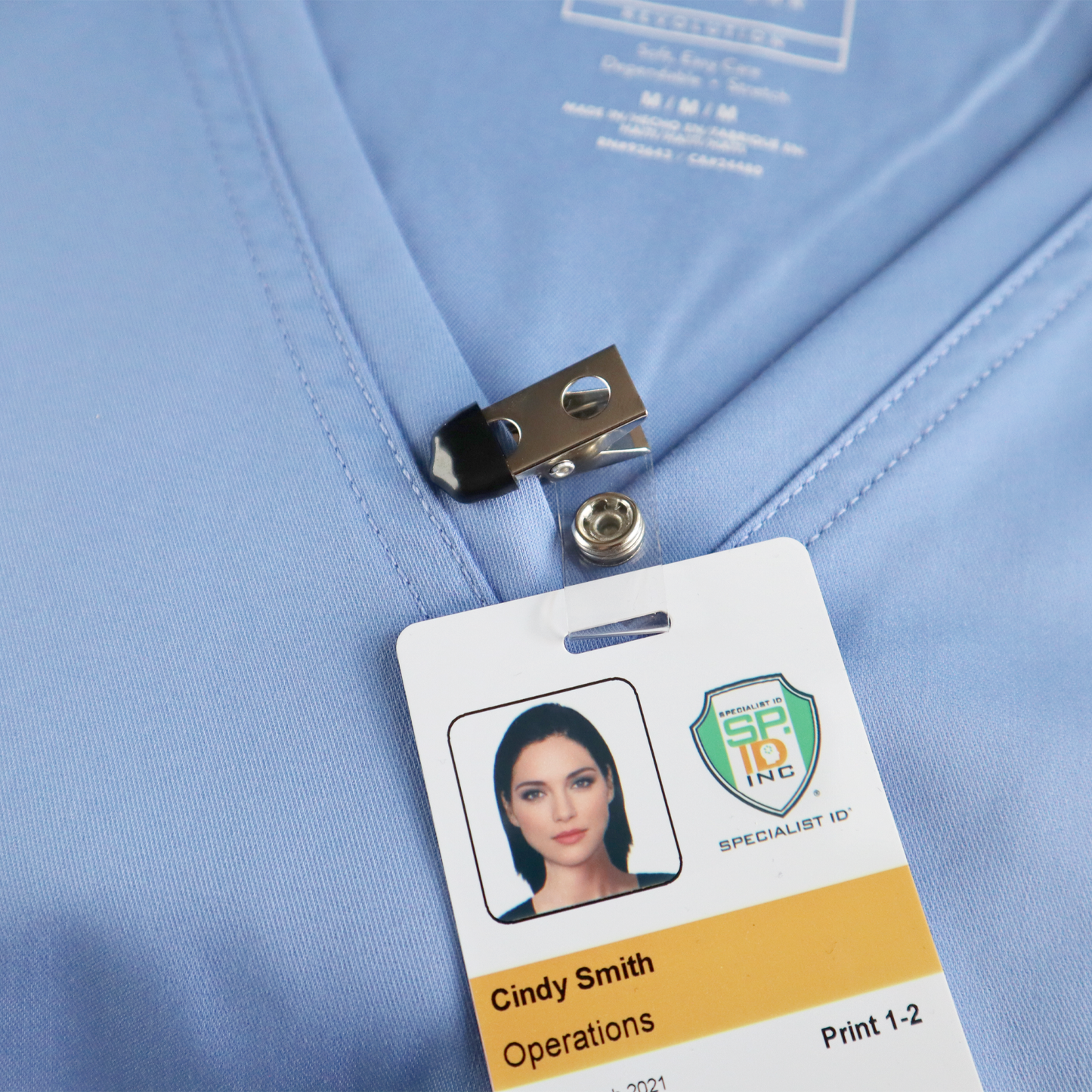 A Clothing Protector ID Strap Clip with Rubber Tip, 2105-3170, secures a name badge to a light blue shirt, displaying a woman's photo, the name "Cindy Smith," and the word "Operations.