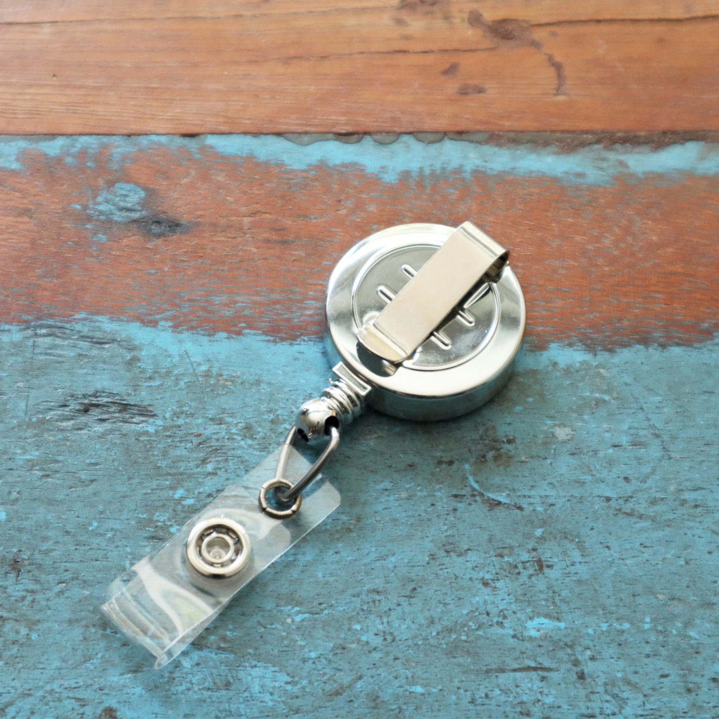 A Silver Chrome Plastic Badge Reel With Belt Clip (P/N 2120-3030) rests on a textured blue and brown wooden surface.