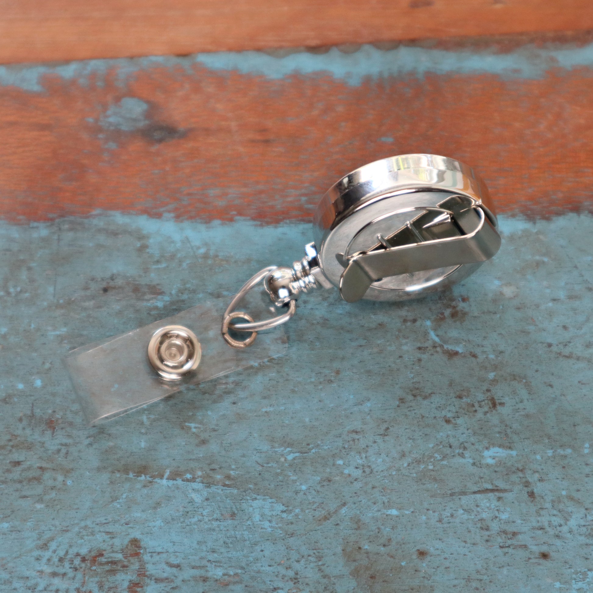 A Silver Chrome Plastic Badge Reel With Belt Clip (P/N 2120-3030) featuring a clear plastic strap is placed on a distressed blue surface.