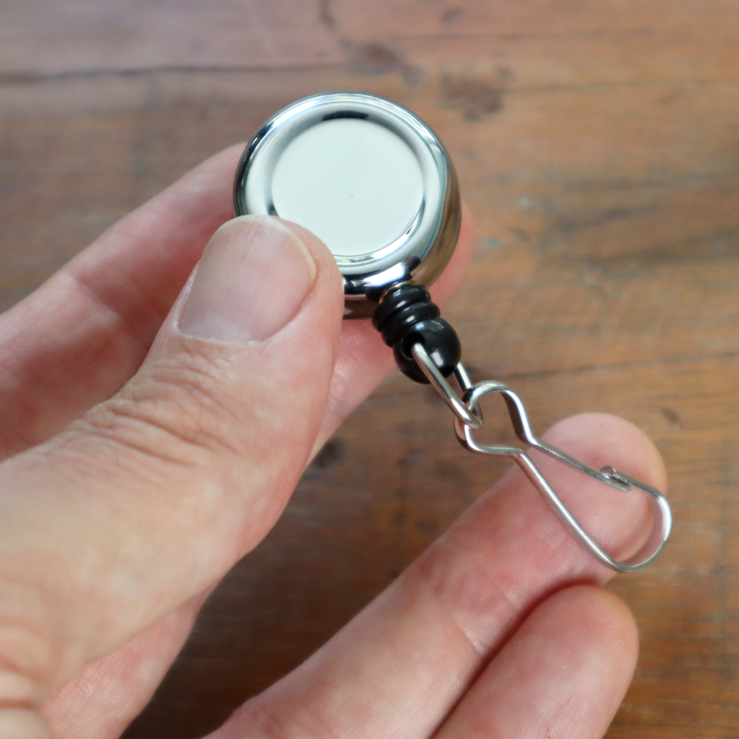 A hand holding a Mini Chrome Silver Key Reel with a spring clip and metal swivel hook against a wooden background.