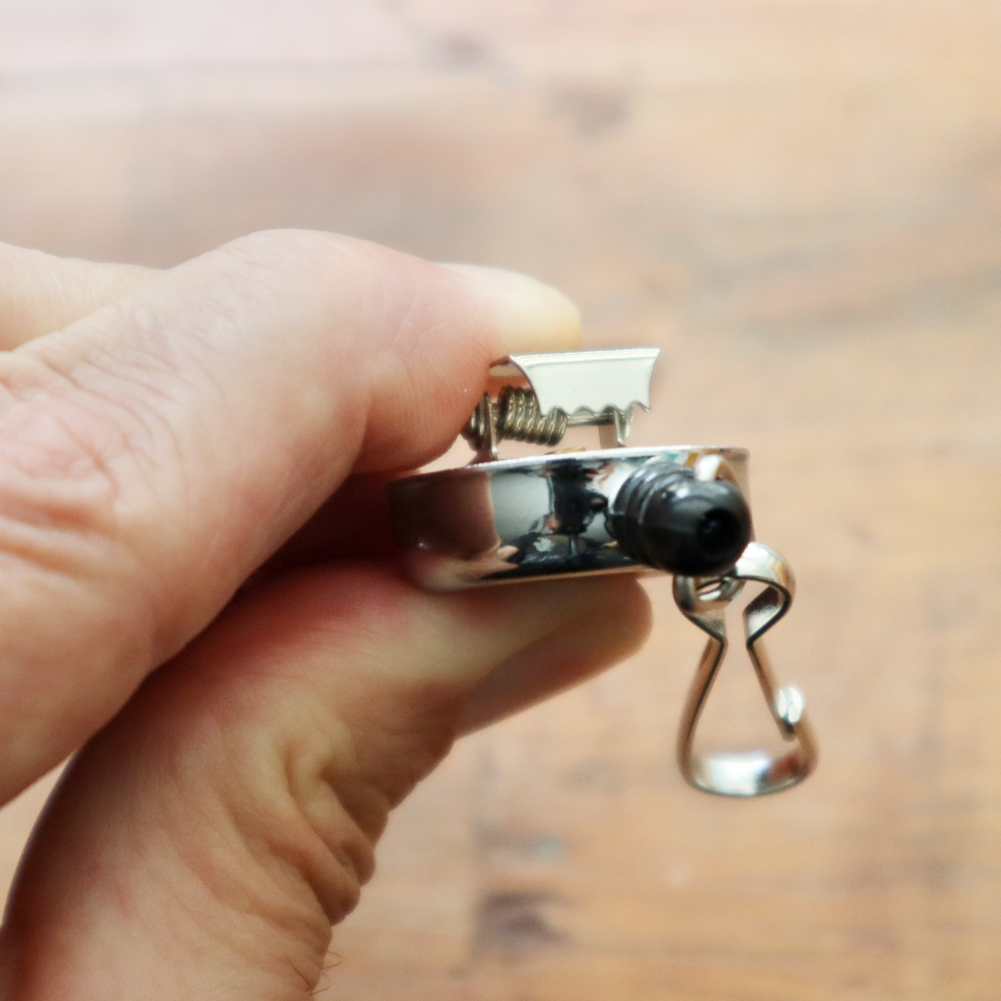 A hand holds a shiny, chrome silver mini key reel with a spring clip and metal swivel hook. The background features a blurred wooden surface, creating an elegant simplicity.