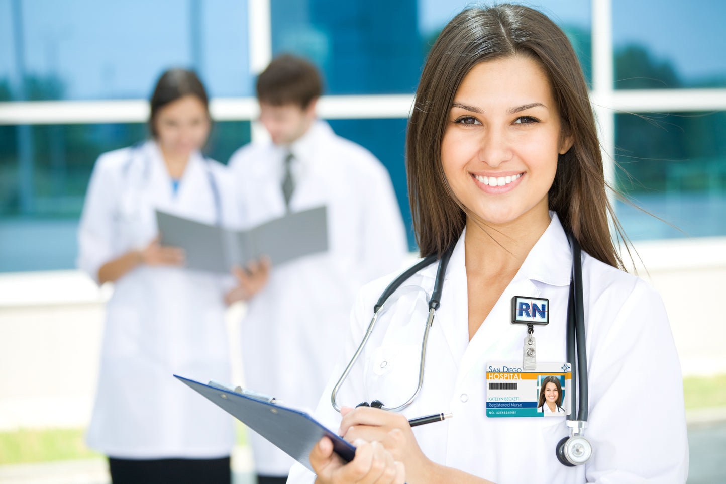 A nurse smiles while holding a clipboard in the foreground, her Custom Rectangle Badge Reel Retractable with Rotating Swivel Clip (P/N 2120-390X-Custom) subtly visible. Two colleagues, one male and one female, review a document in the background.