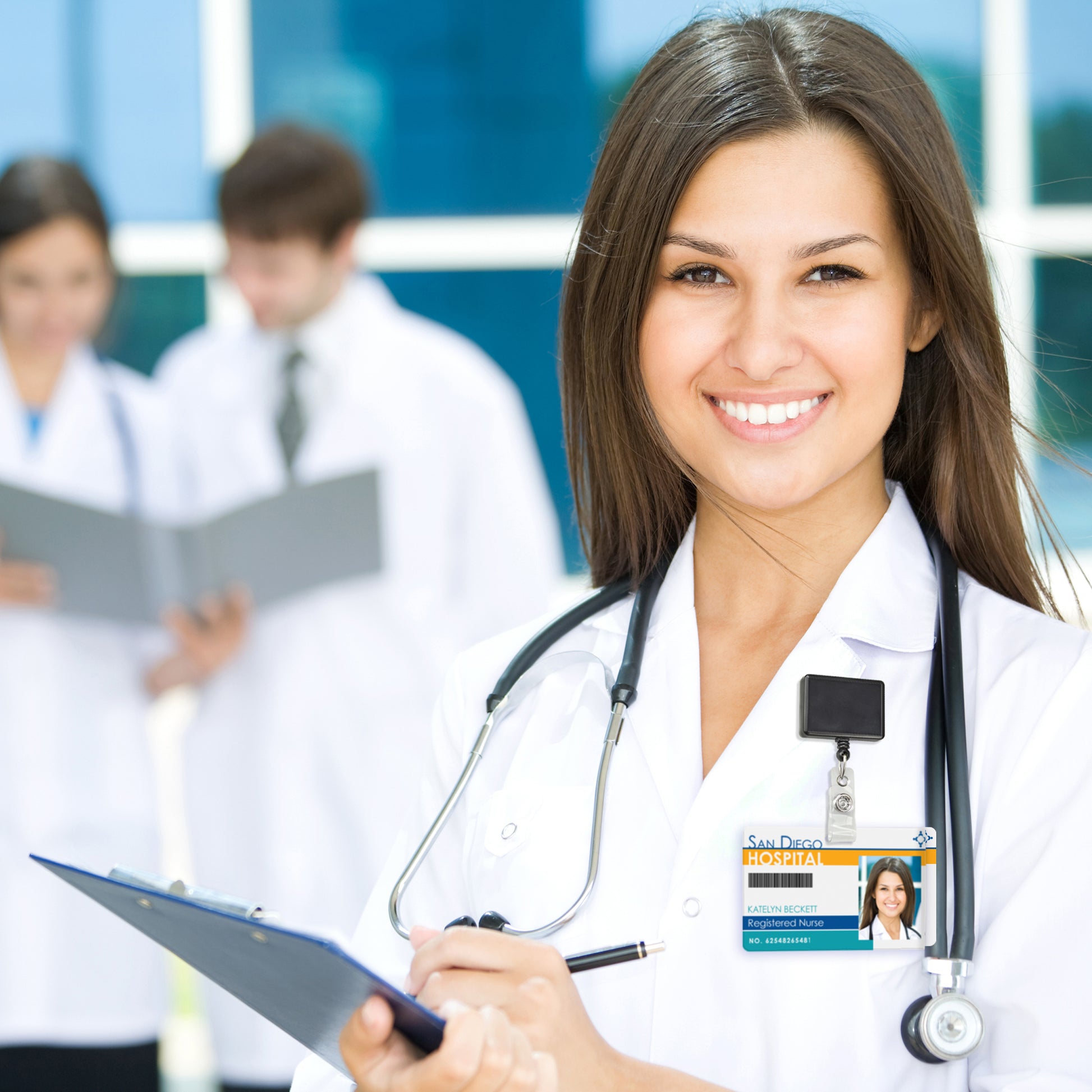 A female doctor smiles at the camera holding a clipboard, with a Rectangle Badge Reel Retractable with Rotating Swivel Clip - Rectangular Badge Holder Reel (2120-390X) attached to her coat. In the background, two other medical professionals are deep in discussion.