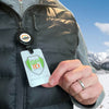 A person wearing a gray zip-up jacket and black vest holds an ID badge lanyard with the 