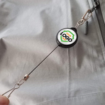 A Custom Retractable Ski Pass Holder - Heavy Duty Sport Reel (2120-4101) - Add Your Logo attached to a zipper on a gray garment, featuring a custom logo on its round casing.