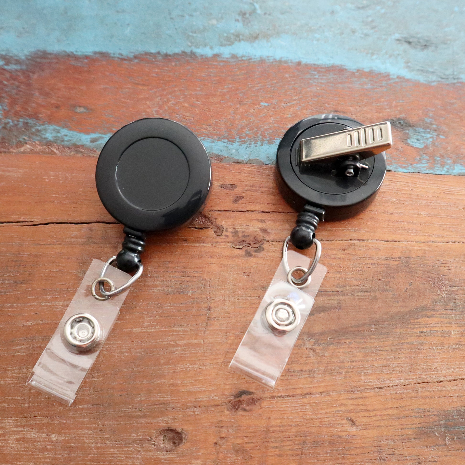 Two Badge Reels with Swivel Spring Clips (P/N 2120-760X) in black, featuring clear plastic card holders, are placed on a wooden surface.