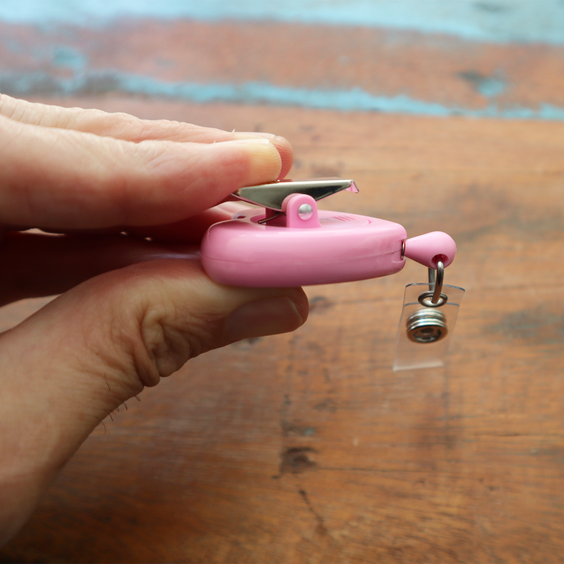 A hand elegantly holds a heart-shaped Ribbon "Awareness" Badge Reel (P/N 2120-7630) with a pink retractable design and a robust metal swivel spring clip, all placed on a wooden surface.