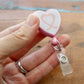 A hand holds the Heart Shaped Ribbon "Awareness" Badge Reel with a pink border and swivel spring clip, displaying an awareness ribbon design on a wooden surface.