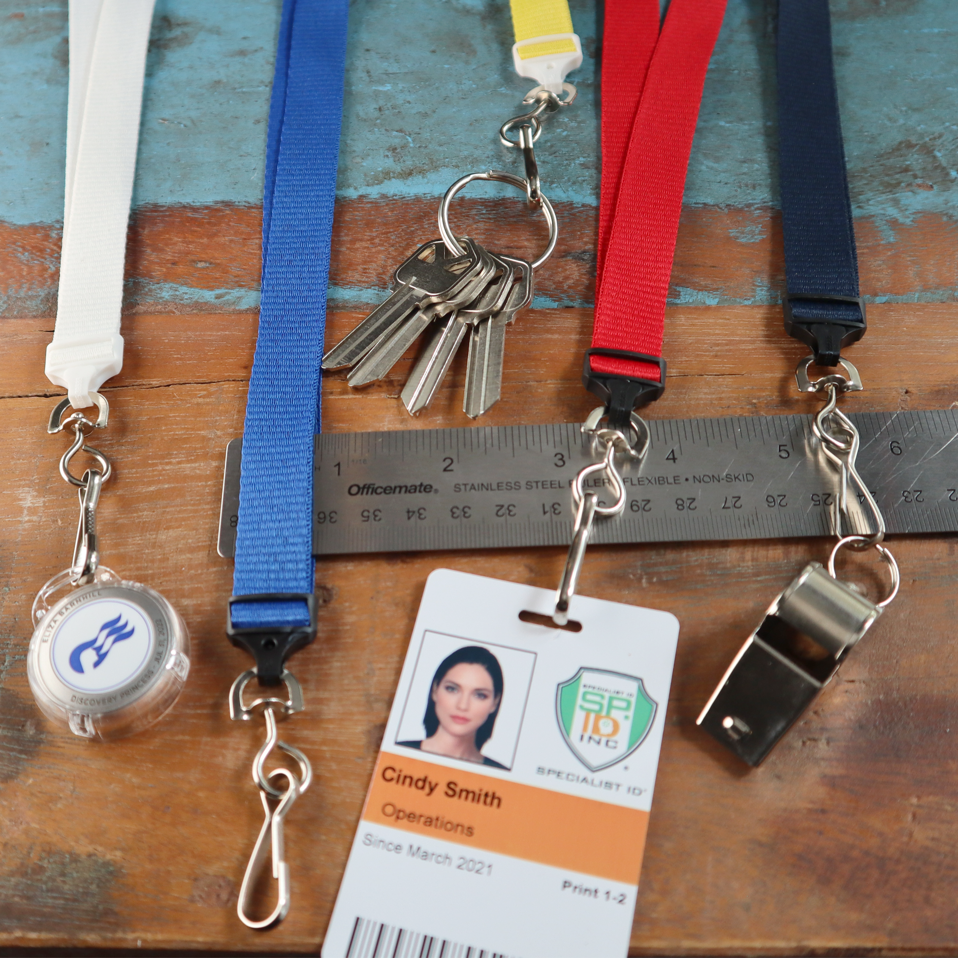 A set of five Flat Wide Breakaway Lanyards with Swivel Hooks (2138-500X) - 5/8 inch width, each featuring different attachments including keys, a retractable badge holder, a whistle, an ID card for Cindy Smith from SF19 Inc., lying on a wooden surface with a ruler.