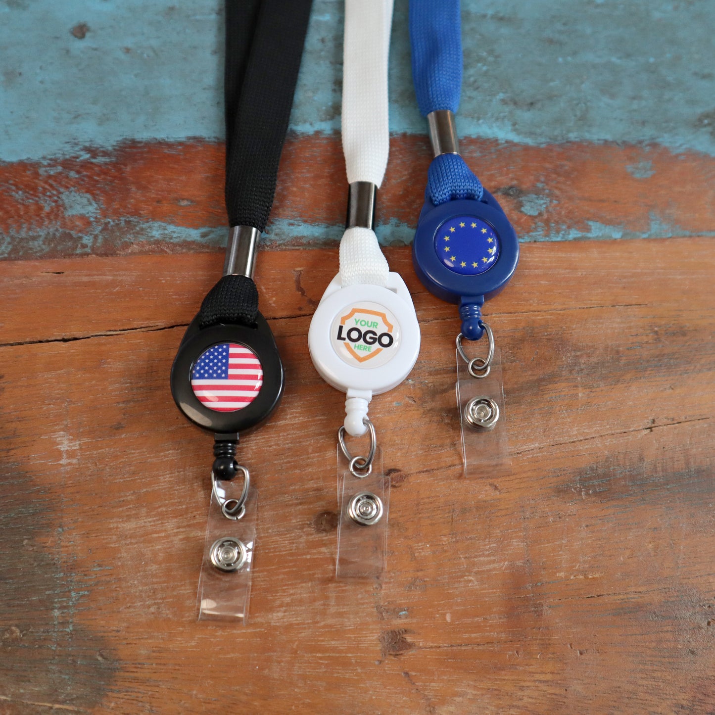 Three lanyards on a wooden surface: a black lanyard with a US flag badge, a white **Breakaway Lanyard with Custom Badge Reel Combo - Add Your Logo to Customize**, and a blue lanyard with an EU flag badge. These Breakaway Lanyards with Custom Badge Reel Combos not only enhance brand awareness but also feature convenient badge reels for easy access.