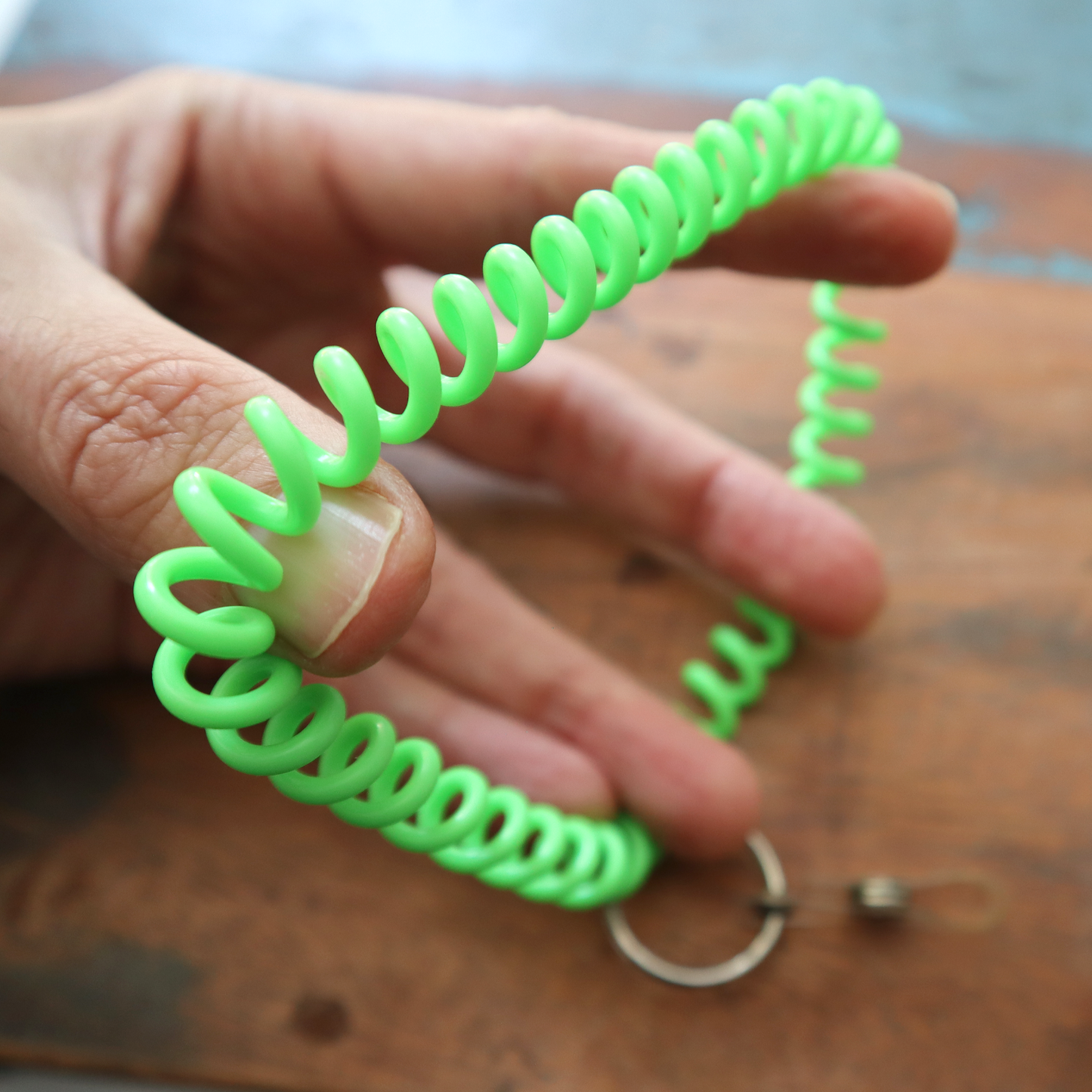 A hand holds the Wrist Coil Key Chain with ID Strap Clip (2140-620X), featuring a bright green coil and a metal ring for secure access against a wooden surface.