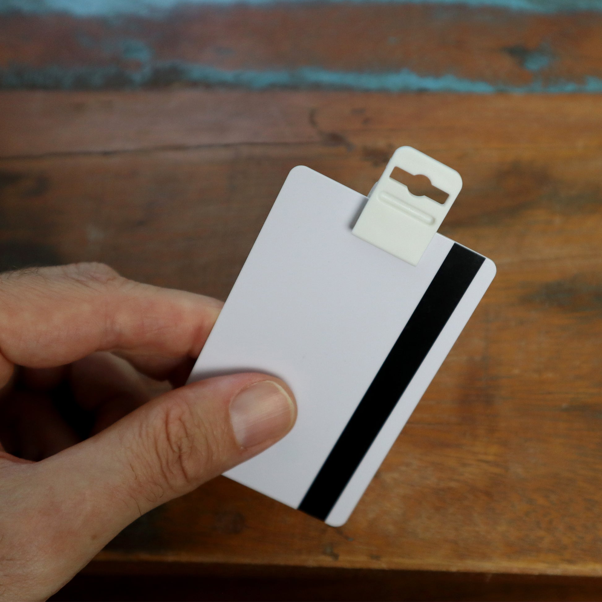 A hand holds a white card equipped with a magnetic stripe, secured by the Gripper 30 Card Clamp (P/N 5710-3058 White), featuring a plastic clip, all resting on a wooden surface.