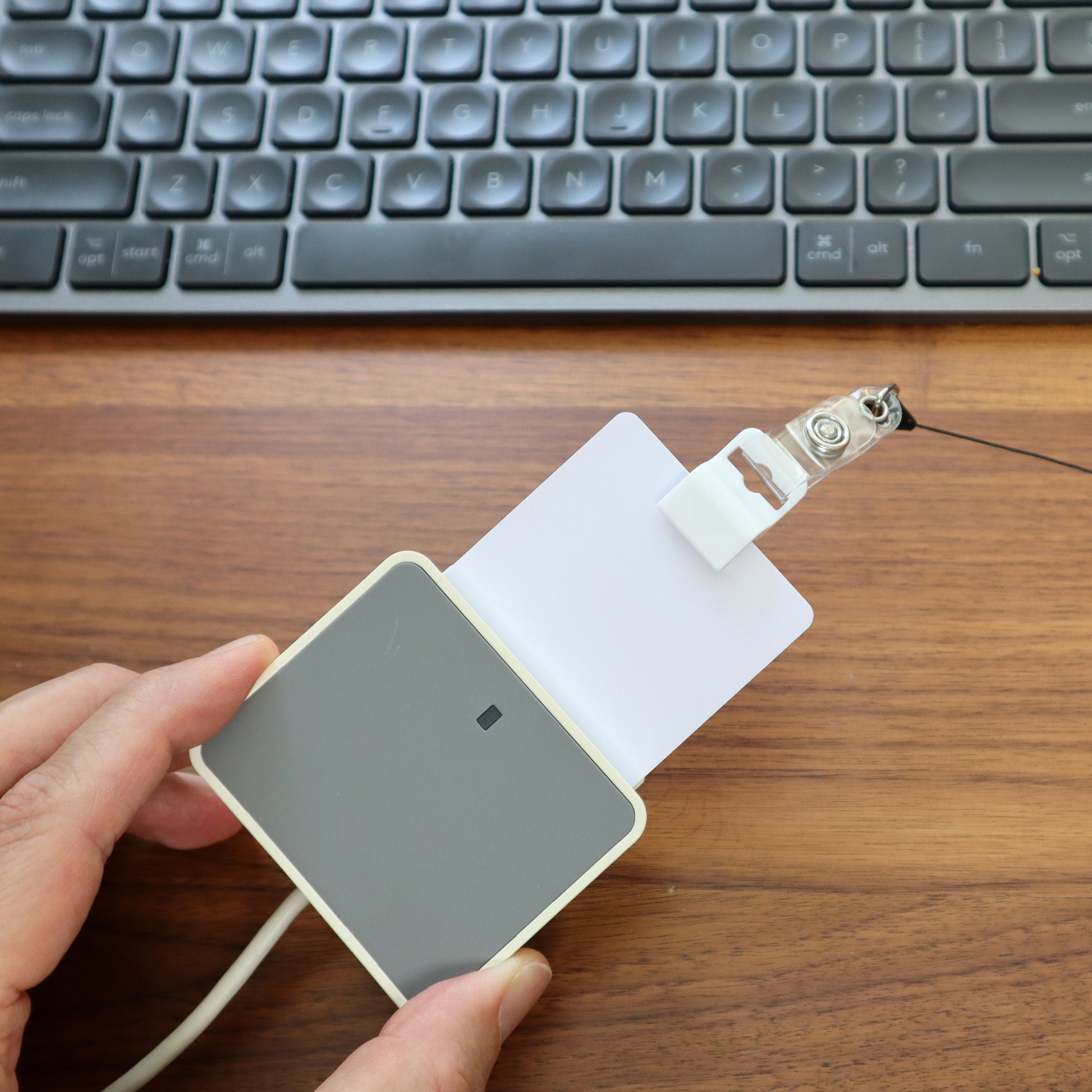 A hand holds an ID card secured by a Gripper 30 Card Clamp (P/N 5710-3050 Black or 5710-3058 White) over a gray card reader on a wooden desk, with a keyboard in the background.