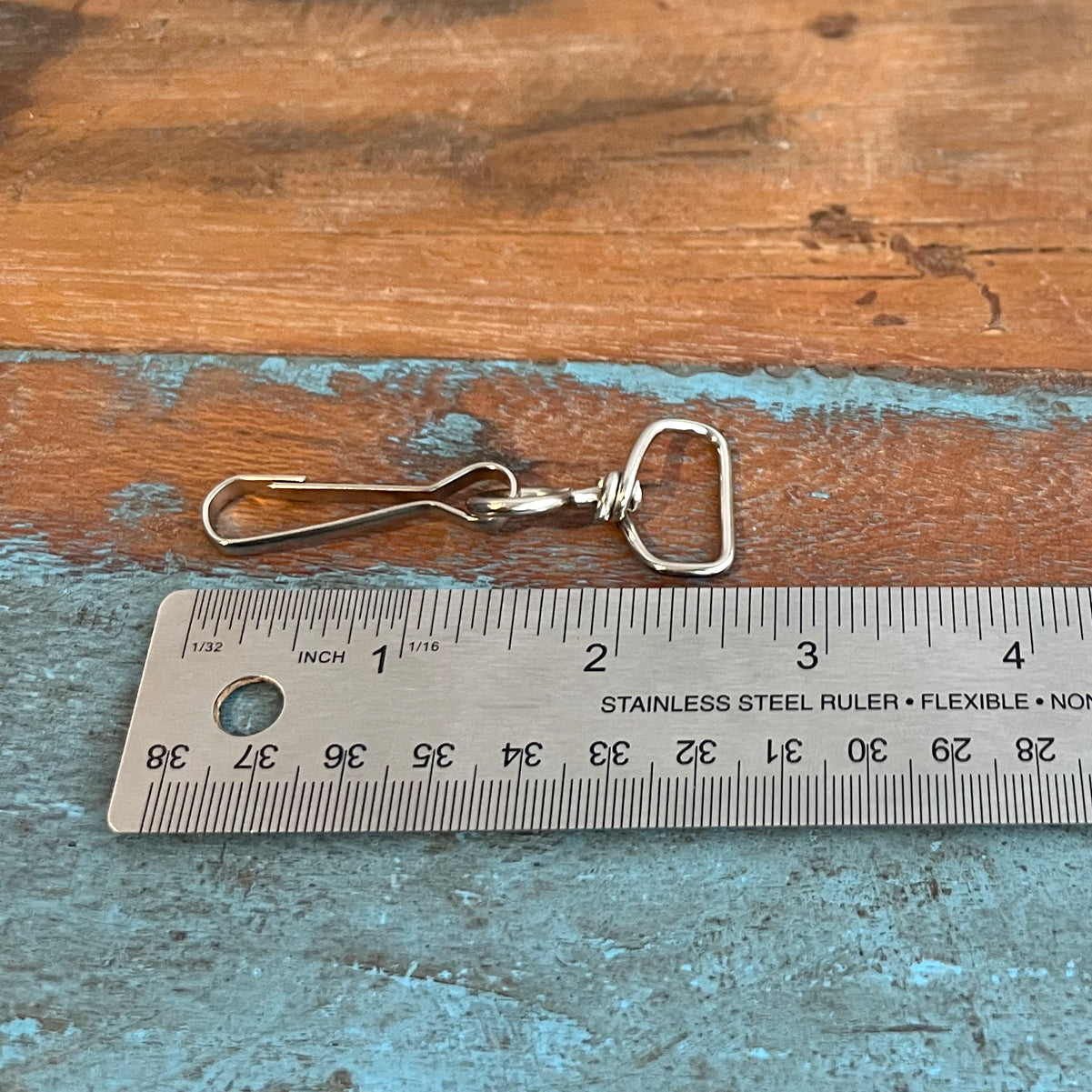 A stainless steel ruler next to a Premium Swivel J Clip Clasp with Wide, Smooth Rounded 3/4 Inch D Ring - DIY Lanyard and Craft Accessories (6905-M-289), placed on a wooden surface. The ruler shows measurements in both inches and centimeters, perfect for precision tasks involving DIY lanyards or swivel J hook clasps.