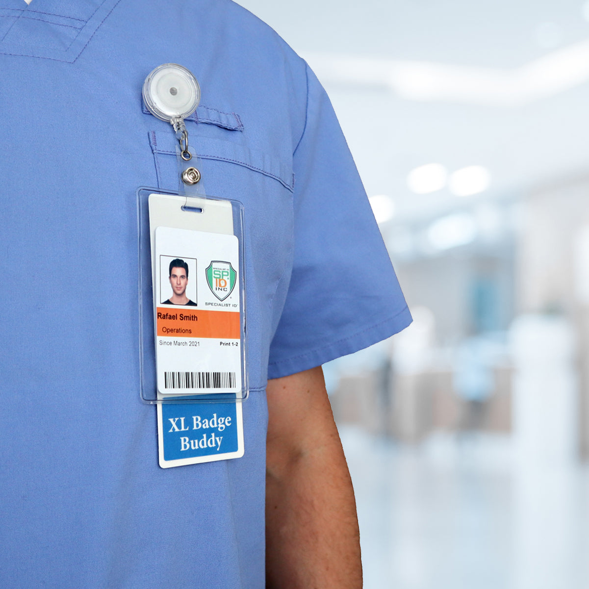 A close-up shows the chest area of a person wearing blue medical scrubs. The visible Oversized NURSE Badge Buddy - Extra Large Badge Buddies for Nurses - Vertical Hospital ID Badge Backer reads "Rafael Smith" and identifies them as an operations staff member. The background is a blurred indoor healthcare setting.