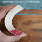 A hand holding a bent plastic label on a wooden surface with the text "Super Durable! We Call Them 'Un-Breakable'" in the background, highlighting the superior quality of our Oversized Custom Vertical Badge Buddy XL- (Extra Large Size).