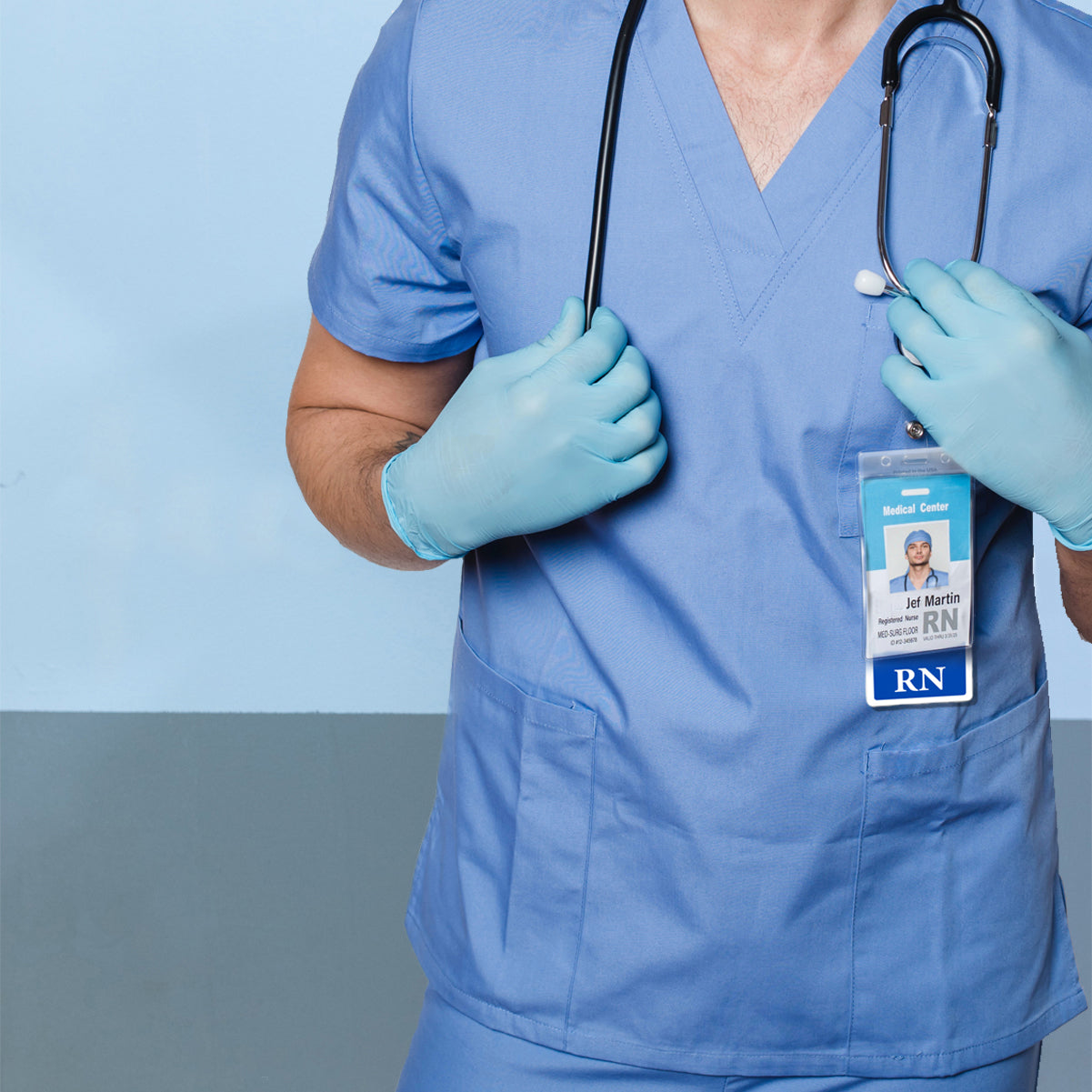 Person wearing blue scrubs, gloves, and a stethoscope around the neck, holding the stethoscope. A visible Oversized RN Badge Buddy - Extra Large Badge Buddies for Registered Nurse - Vertical Hospital ID Badge Backer prominently displays their name and designation as a registered nurse (RN).