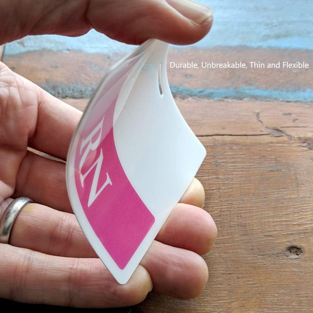 A hand bends a flexible, thin card labeled "RN" in pink, resembling an MD Horizontal Badge Buddy with a RED Border against a wooden surface.