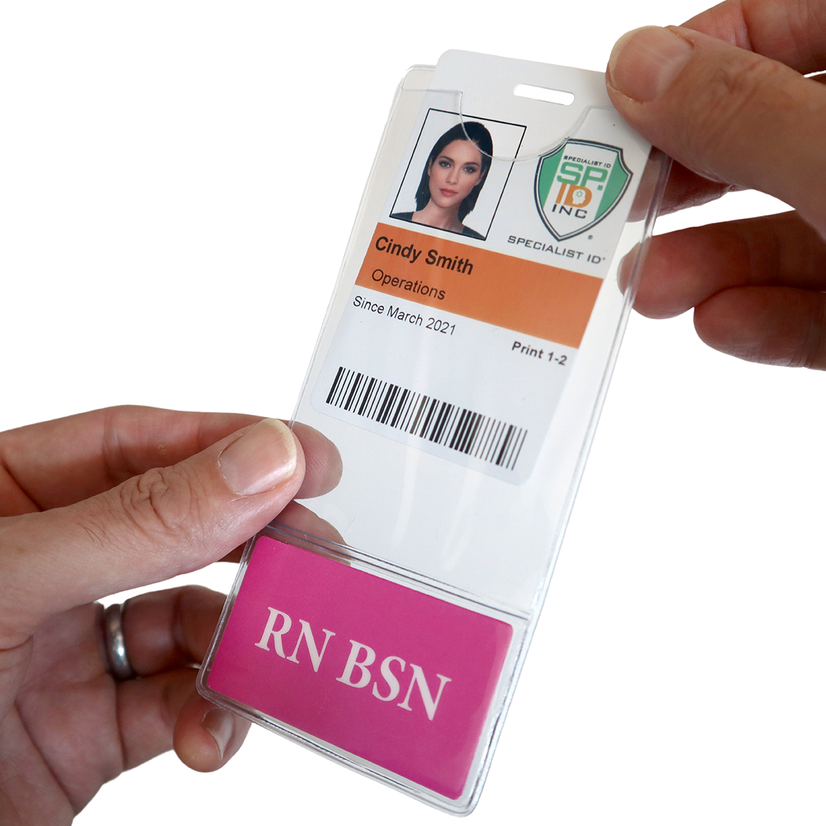 Hands holding an ID badge with a photo, name (Cindy Smith), and title (Operations), indicating employment since March 2021 and a bar code. Below is a pink card with "RN BSN." The ID badge is securely placed in an RN BSN BadgeBottom Badge Holder & Badge Buddy IN ONE!! - Vertical ID Badge Sleeve with Bottom Role Tag for Registered Nurses.