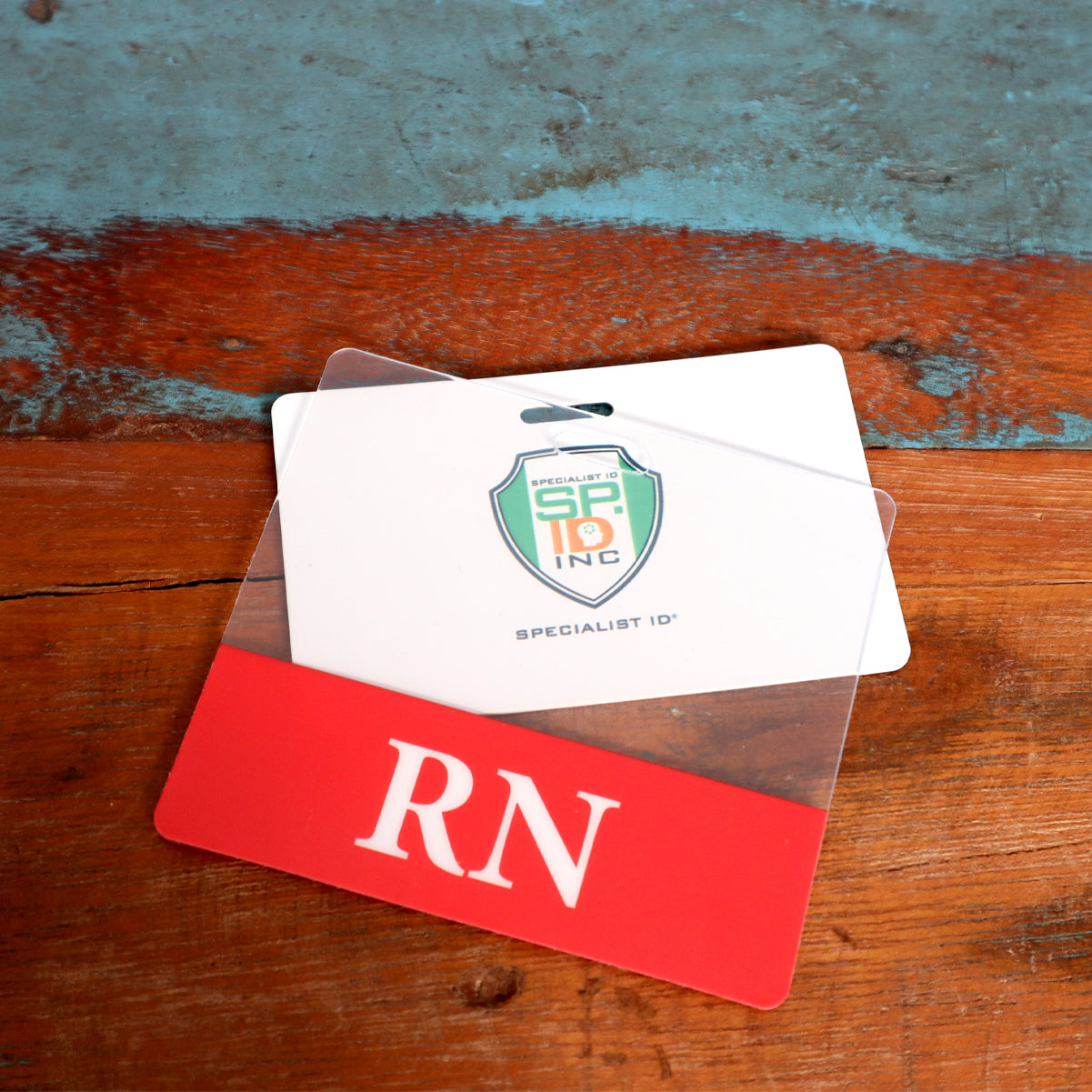 A white ID badge with a green logo and the text "Specialist ID" sits on a wooden surface. A Clear RN Badge Buddy - Horizontal ID Badge Backer for Nurses - Double Sided Print is placed over part of the badge, creating an efficient and professional display.