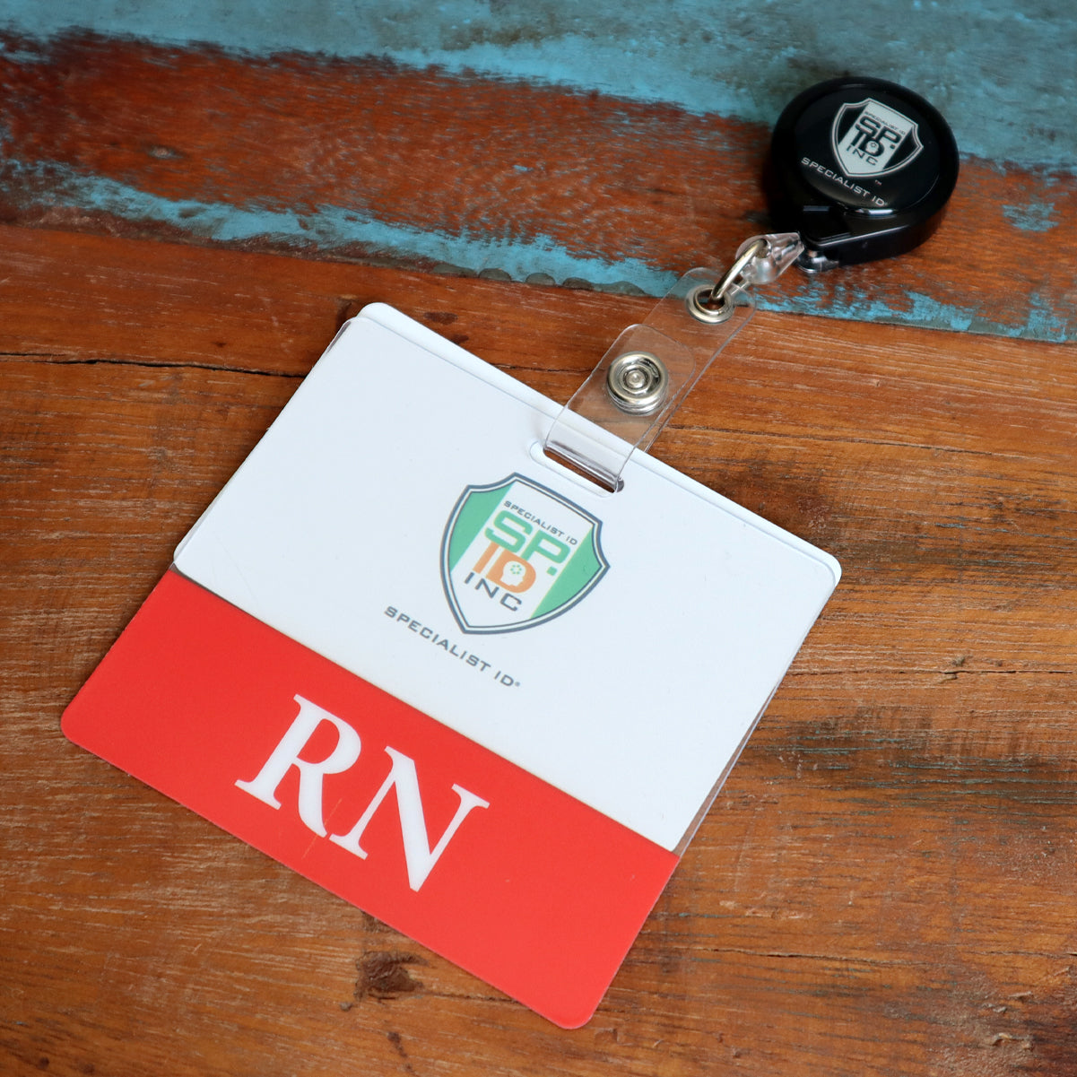 A name badge holder on a retractable clip is placed on a wooden surface. The Clear RN Badge Buddy - Horizontal ID Badge Backer for Nurses - Double Sided Print has an emblem and the letters "RN" printed on it.