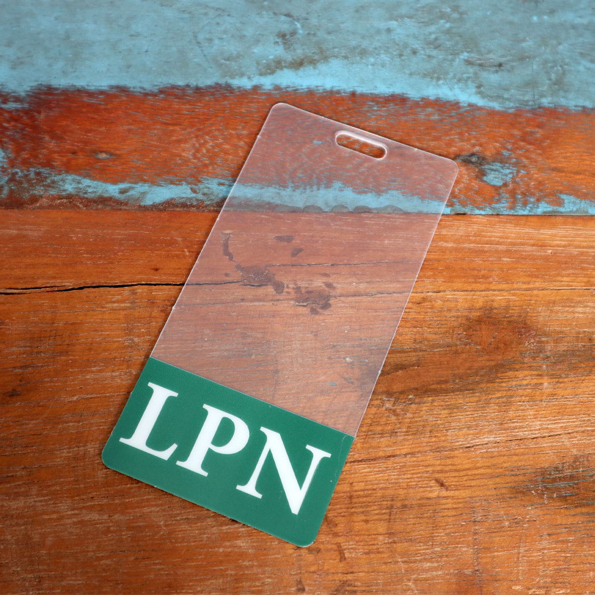 A **Clear LPN Badge Buddy Vertical with Green Border for Licensed Practical Nurses** is placed on a wooden surface, functioning as an LPN Badge Buddy for Licensed Practical Nurses.