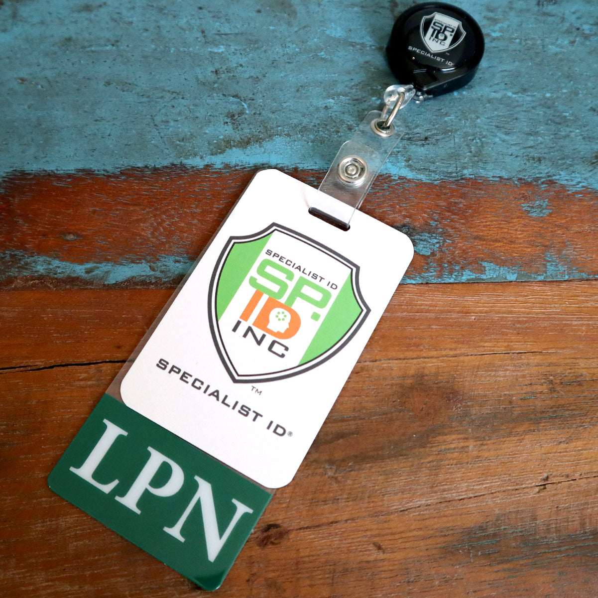 An LPN badge with a "Specialist ID Inc." logo on a retractable badge holder and an additional Clear LPN Badge Buddy Vertical with Green Border for Licensed Practical Nurses rests on a wooden surface.