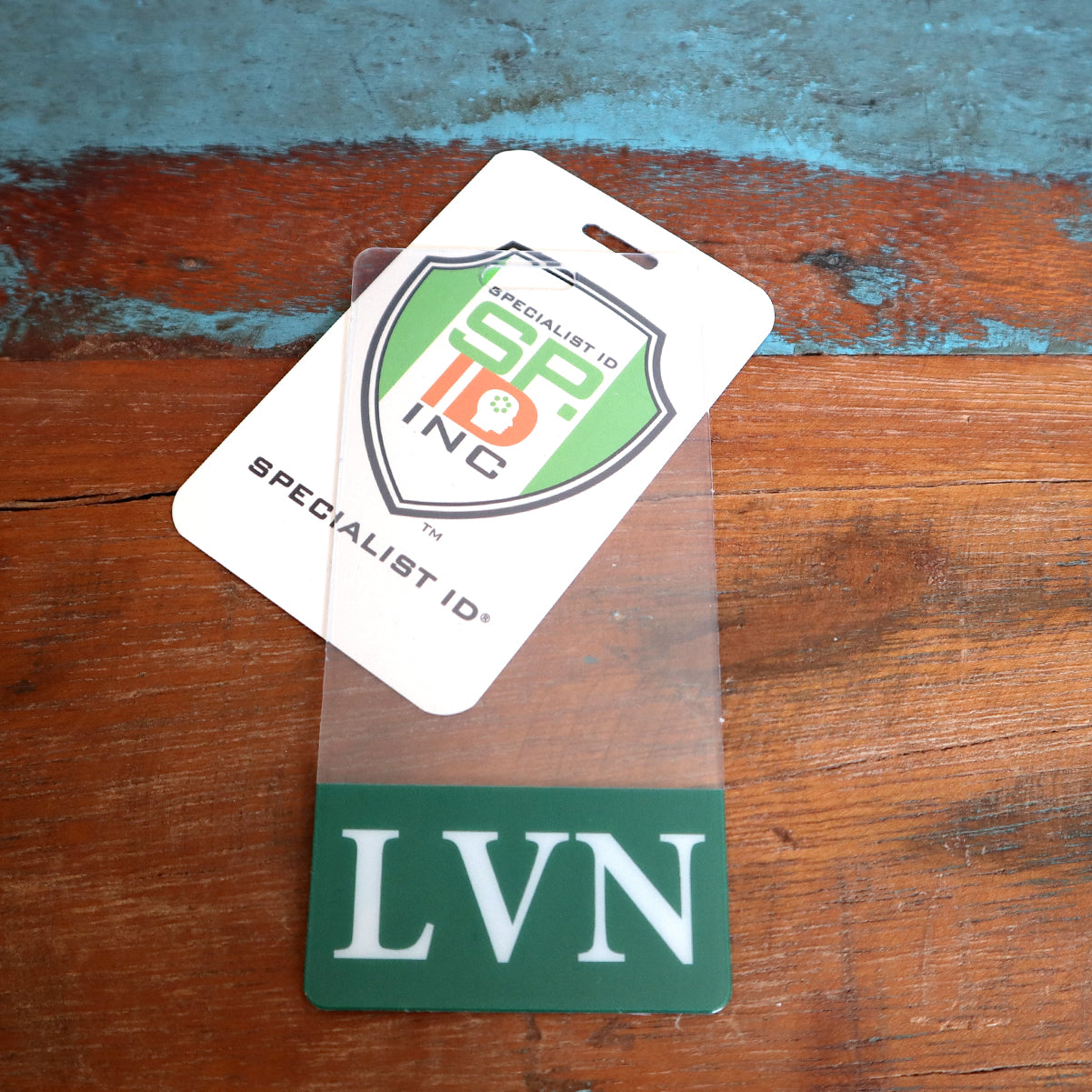 A **Clear LVN Badge Buddy Vertical with Green Border for Licensed Vocational Nurses** lies on a wooden surface next to a Specialist ID card, showcasing a vertical ID badge for licensed vocational nurses.