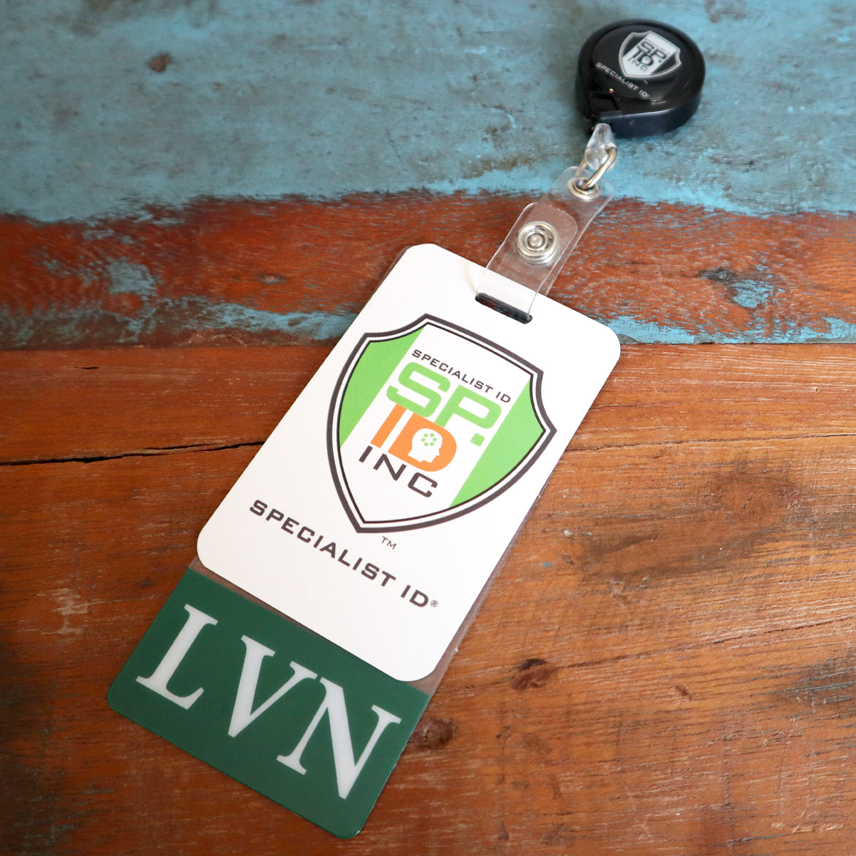 An ID badge with the logo "Specialist ID Inc." and acronym "LVN" at the bottom is clipped to a retractable badge reel. The Clear LVN Badge Buddy Vertical with Green Border for Licensed Vocational Nurses is placed on a worn, blue-painted wooden surface.