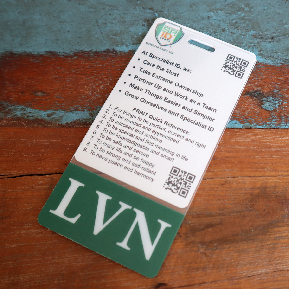 An identification card displayed on a textured surface. The vertical ID badge features a list under the "Specialist ID" heading, with "LVN" at the bottom. There are QR codes and multiple bullet points on the card, making it clear that this is a Clear LVN Badge Buddy Vertical with Green Border for Licensed Vocational Nurses.