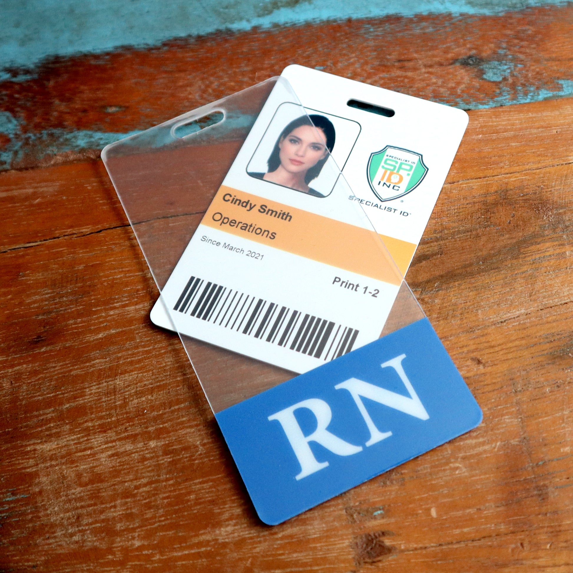 A Clear RN Badge Buddy Vertical with Color Border for Registered Nurses - Double Sided Print, customized for Cindy Smith in Operations, featuring her photo and barcode along with the label "RN," is placed on a wooden surface for instant role recognition.