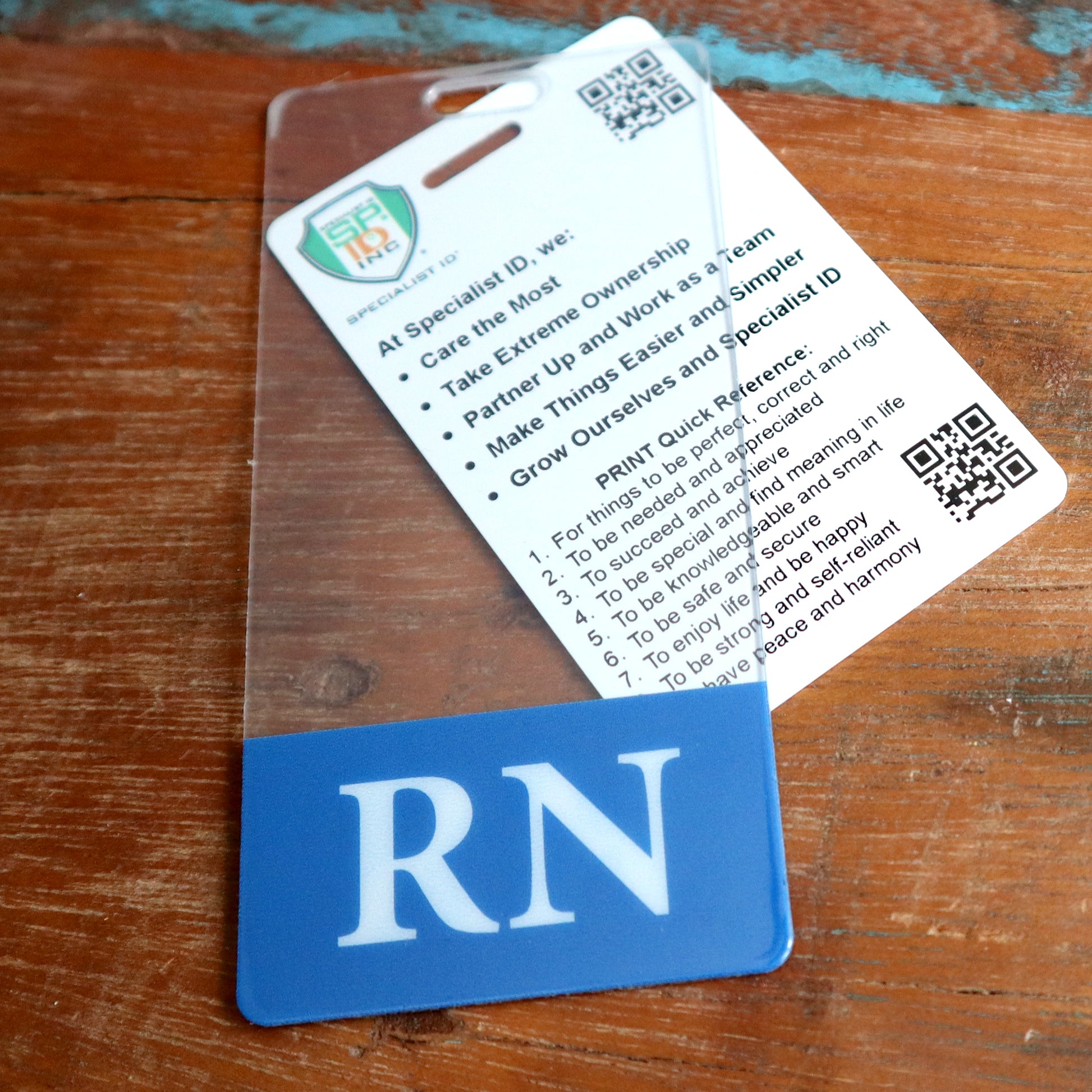 A Clear RN Badge Buddy Vertical with a color border for Registered Nurses lies on a wooden surface. The double-sided ID backer contains a card with printed text and QR codes, listing responsibilities and commitments related to the professional role, ensuring instant role recognition.