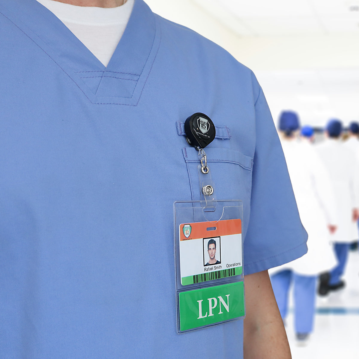 Person wearing blue medical scrubs with an LPN badge held securely in the LPN BadgeBottom Badge Holder & LPN Badge Buddy IN ONE!! - Horizontal ID Badge Sleeve with Bottom Role Tag for Nurses clipped to the chest pocket; a group of healthcare professionals is visible in the background.