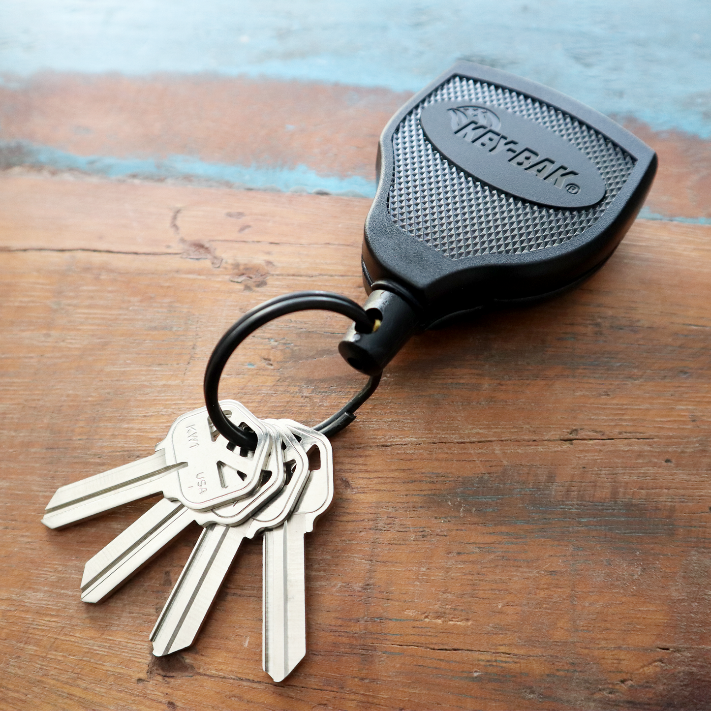 The Key-Bak Super 48 Heavy Duty Key Reel with Belt Clip (S48K), featuring a durable Kevlar cord, displays four keys on a wooden surface, each attached to a keyring with a black key fob.