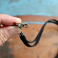 A hand holds a Flat Braid Woven Non-Breakaway Lanyard With a Steel Bulldog Clip (P/N 2135-355X) attached to a black lanyard, with a blurred background of blue and brown surfaces.