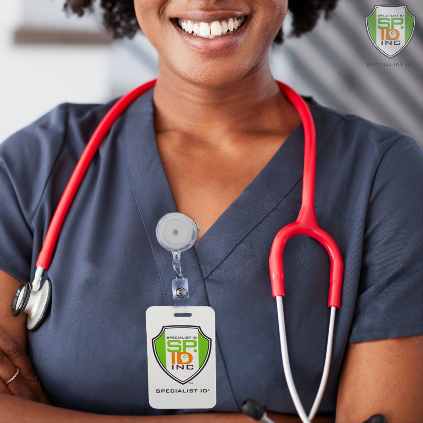 A healthcare worker wearing a blue uniform, a red stethoscope, and an ID badge labeled "Specialist ID Inc" is smiling with arms crossed, with their Translucent Badge Reel with Swivel Clip (P/N 2120-762X) slightly visible.