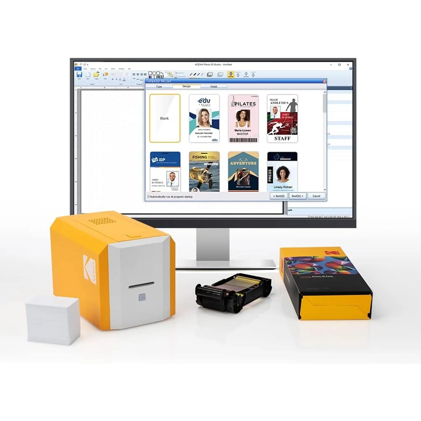 A desktop ID card printing setup featuring a computer monitor displaying card design software, a Kodak ID100S Single-Sided Photo ID Card Printer (Manual Hand-Fed) in yellow and white, blank cards, a card printer ribbon, and a box of customized employee IDs.