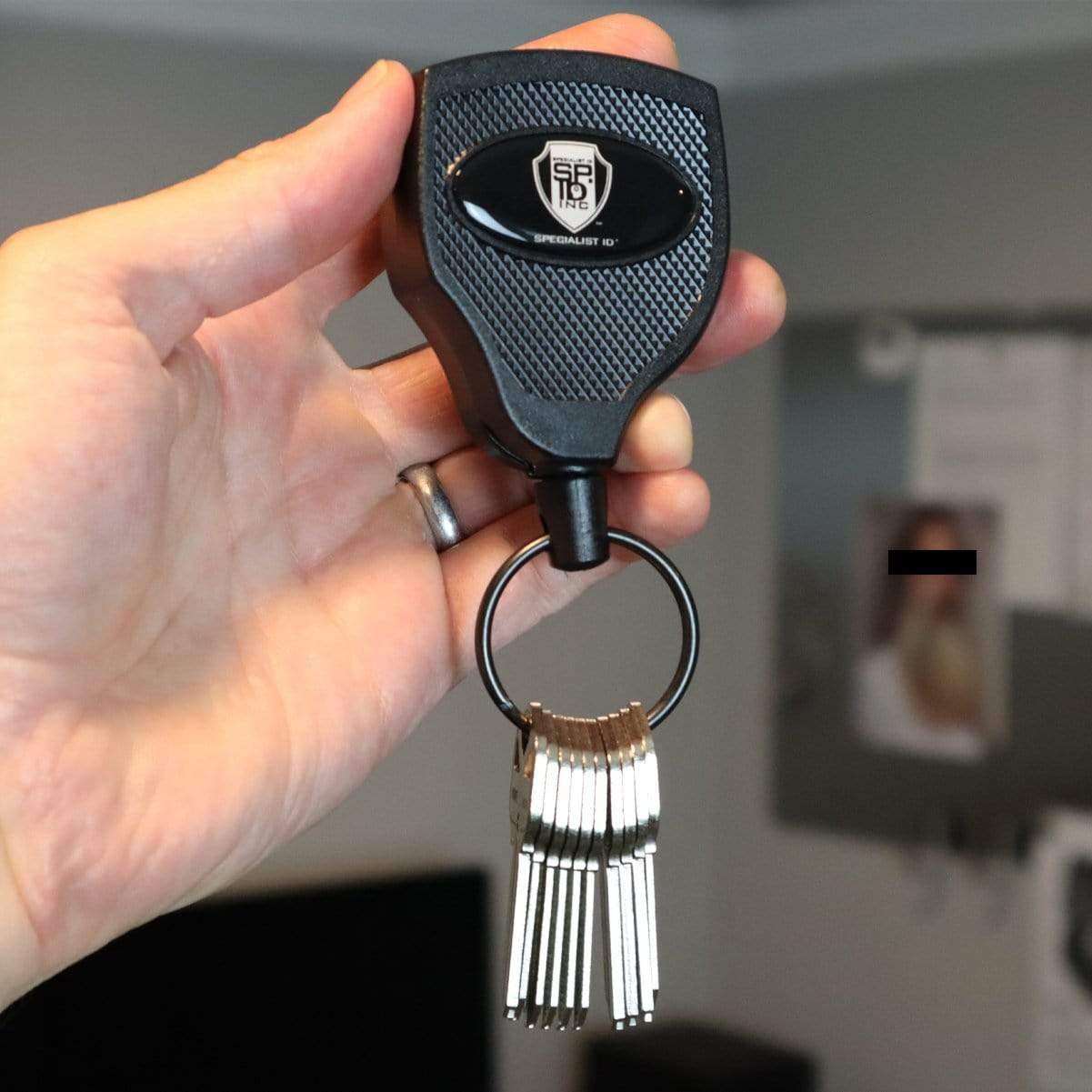 A hand holds a set of keys attached to a Super Heavy Duty Retractable Keychain - 8oz or 10 Keys - Durable 48” (4 Ft) Kevlar Lanyard, featuring an emblem. The background is slightly blurred, showing indistinct objects.