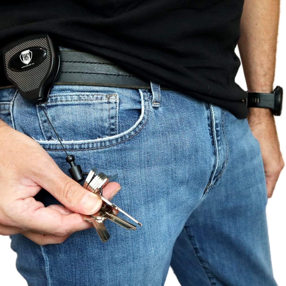 Person wearing a black shirt and blue jeans, holding a set of keys attached to a Super Heavy Duty Retractable Keychain - 8oz or 10 Keys - Durable 48” (4 Ft) Kevlar Lanyard clipped to their waistband.