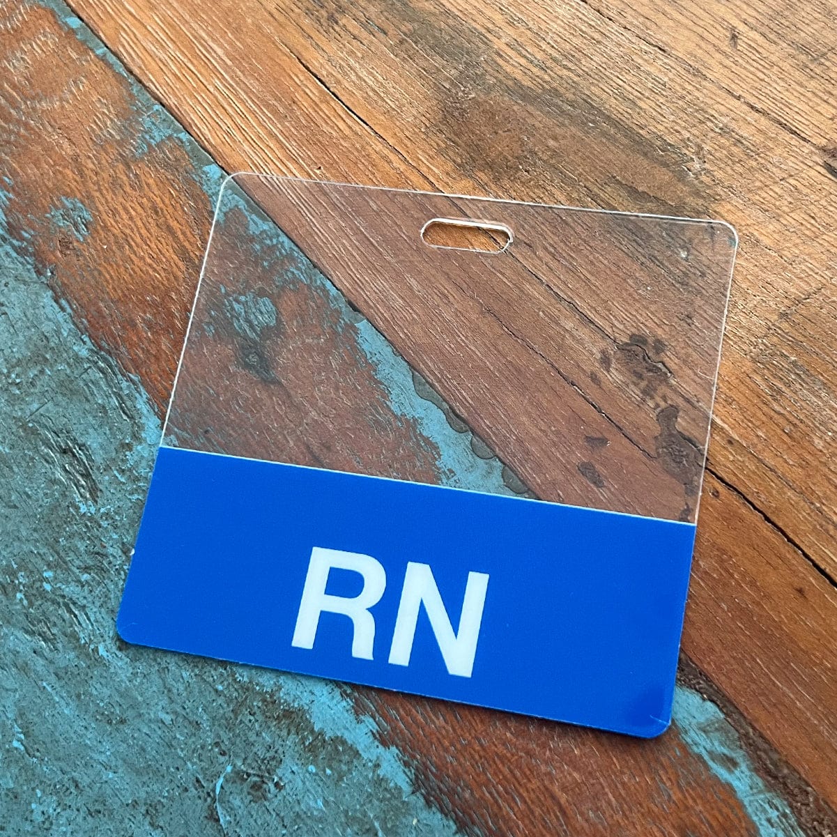 A Clear RN Badge Buddy - Horizontal ID Badge Backer for Nurses - Double Sided Print with a blue bottom, featuring the letters "RN" printed in white, serves as an ideal RN Badge Buddy on a wooden and blue surface.