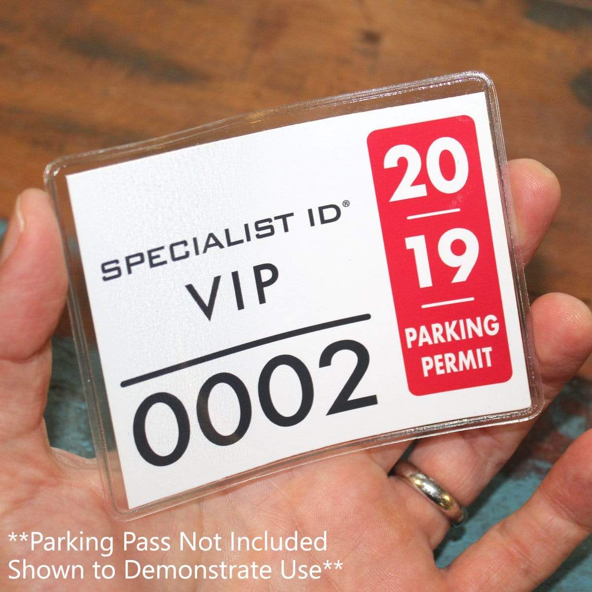 Hand holding a Large 4 x 3 Adhesive Badge Holders with Sticky Back - Clear Vinyl Parking Pass Holders for Windshield (CE-4P) displaying a VIP Specialist ID card with a 2019 parking permit. Text on the image states: "Parking Pass Not Included Shown to Demonstrate Use".