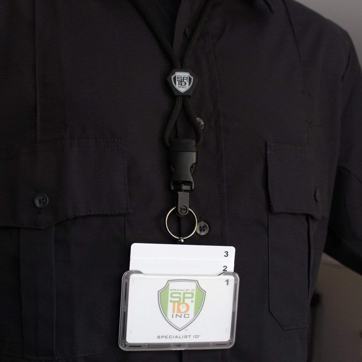 A person in professional work wear is sporting a black shirt with a Specialist ID Horizontal 3 Card Badge Holder & Heavy Duty Lanyard with Breakaway Clip and Key Ring - Hard Plastic Rigid Name Tag Protector - Top Load for Three Badges from "Specialist ID" around their neck. The badge holder contains a name badge and two additional cards.