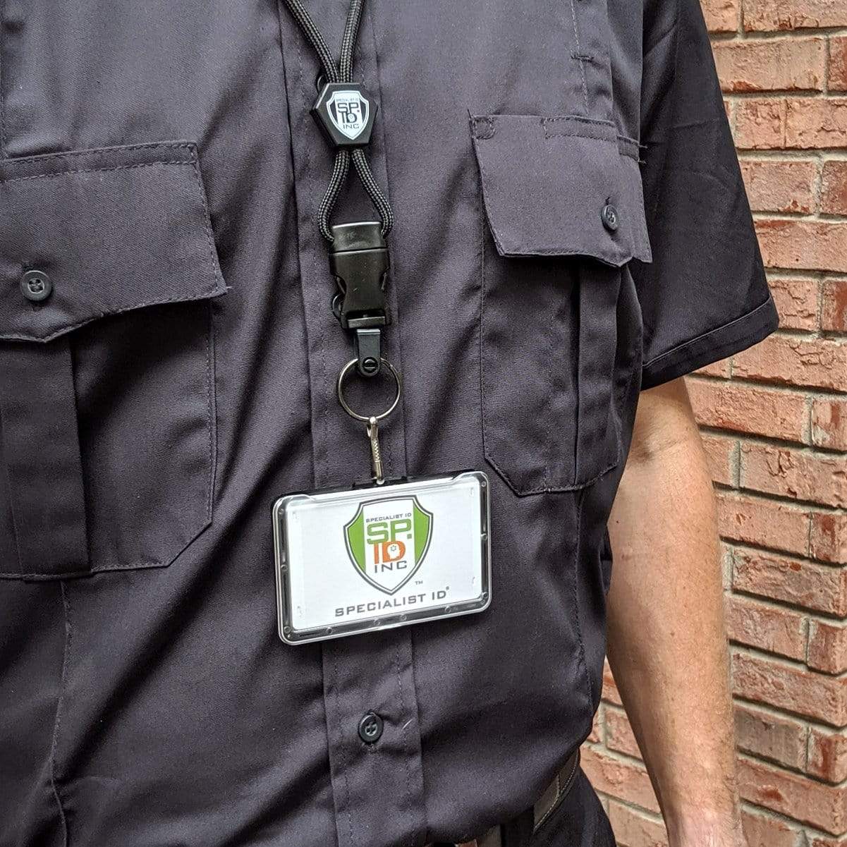 A person standing against a brick wall is wearing professional work wear: a black uniform with dual chest pockets, and displaying a Specialist ID Horizontal 3 Card Badge Holder & Heavy Duty Lanyard with Breakaway Clip and Key Ring - Hard Plastic Rigid Name Tag Protector - Top Load for Three Badges secured in a badge holder hanging from a breakaway lanyard.