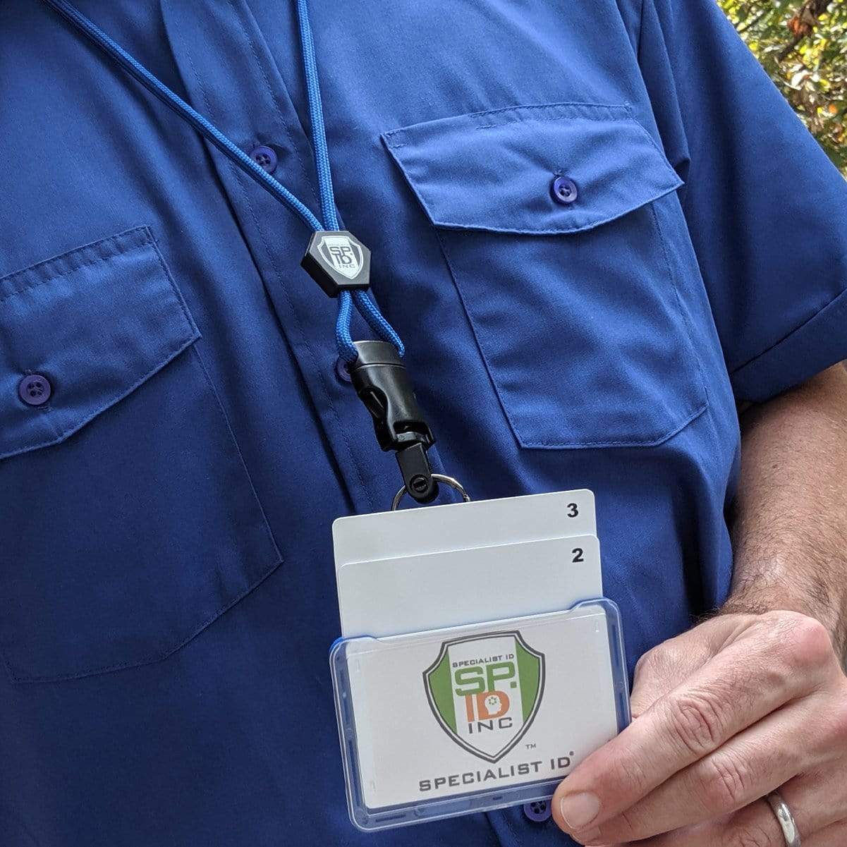 A person wearing professional work wear, a blue shirt with two chest pockets, holds an ID badge on a Specialist ID Horizontal 3 Card Badge Holder & Heavy Duty Lanyard with Breakaway Clip and Key Ring - Hard Plastic Rigid Name Tag Protector - Top Load for Three Badges around their neck. The badge shows "Specialist ID Inc." and the individual has a stack of cards labeled 2 and 3.